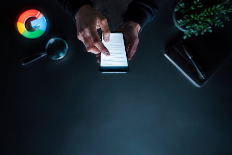 Mains utilisant un smartphone dans l'obscurité avec un logo Google, une loupe et un carnet sur le bureau.